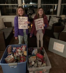 Two young girls standing by two bins full of food and holding signs that read " Foundry Church Food Drive"